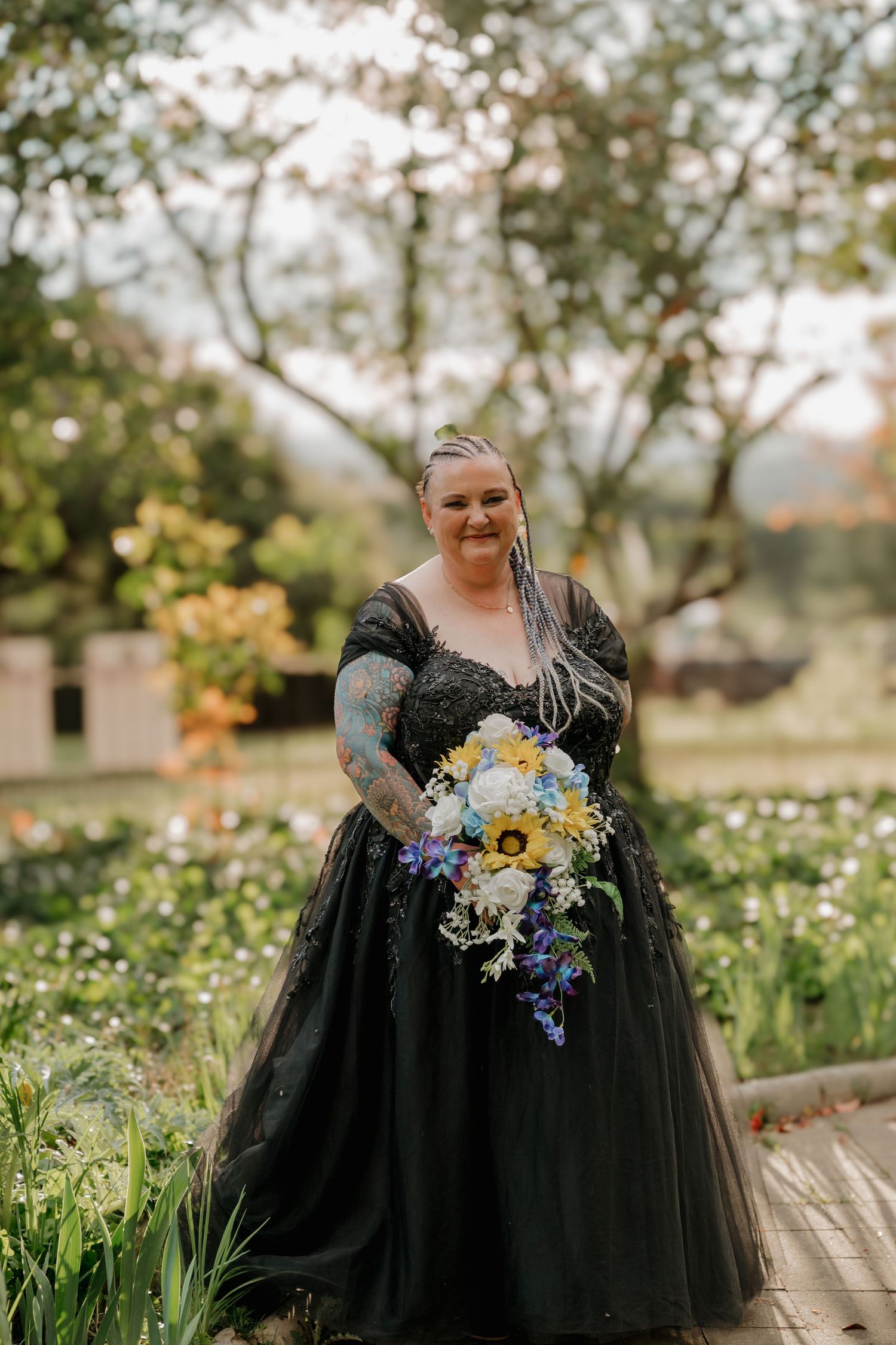 Bride in black ball gown with tattoos holds colorful bouquet with sunflowers in lush garden setting.