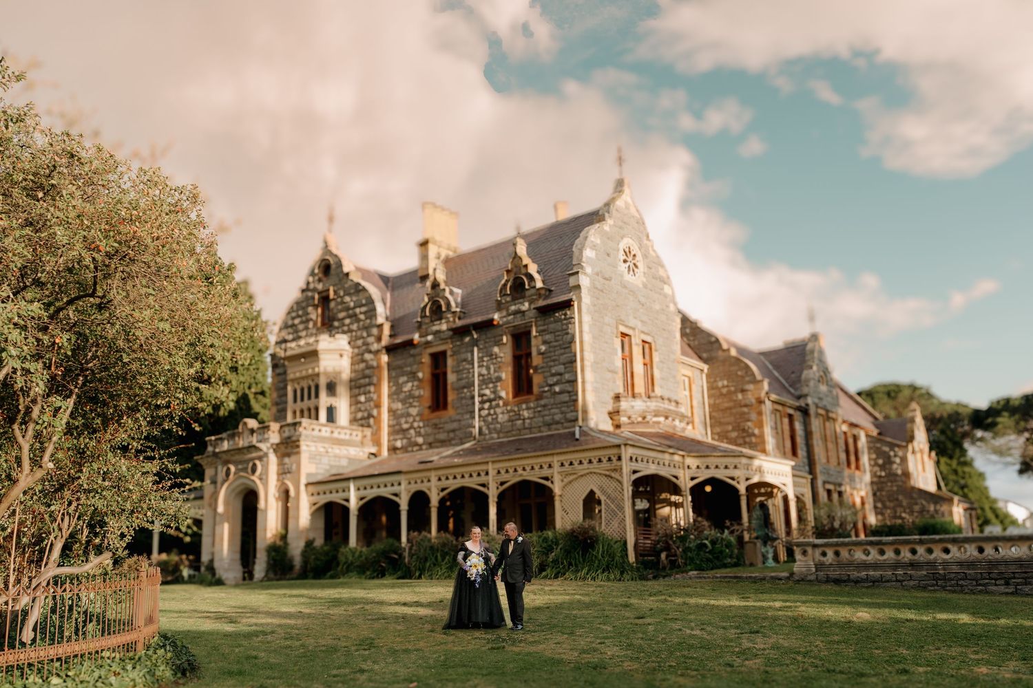 Couple posing in front of a grand historic stone mansion with Victorian architecture on a sunny day.