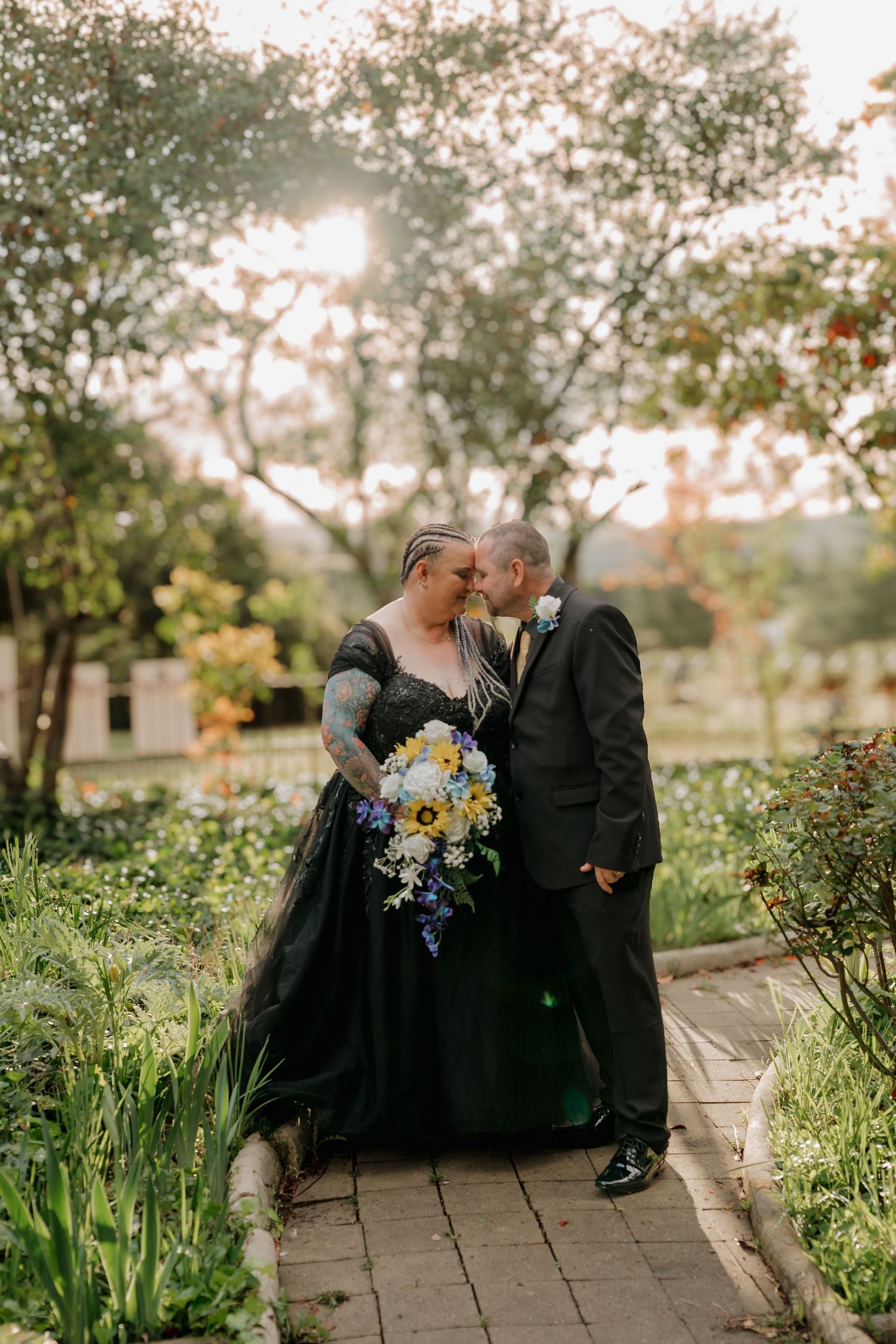 Couple shares romantic moment in garden, bride in black gown with floral tattoos holding sunflower bouquet, groom in black suit.