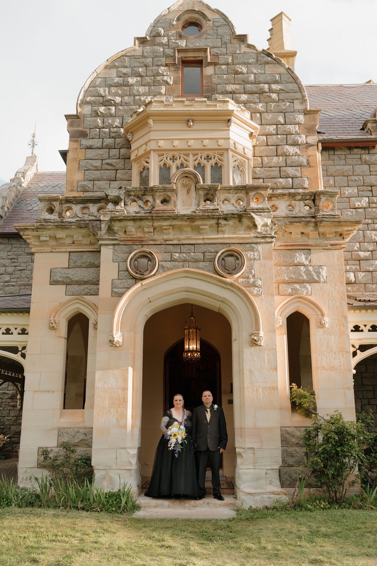 Couple in formal attire posing at ornate stone castle entrance with arched doorway and Gothic architectural details.