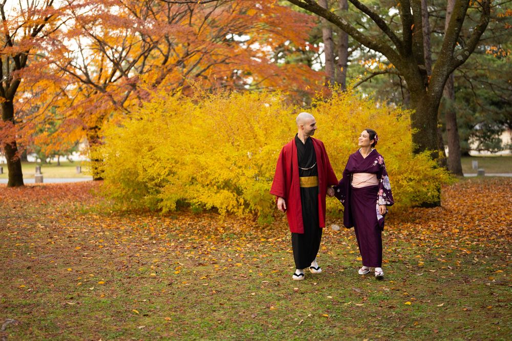 Couple in traditional Japanese kimono holding hands and walking through a park with bright yellow shrubs and orange autumn foliage.
