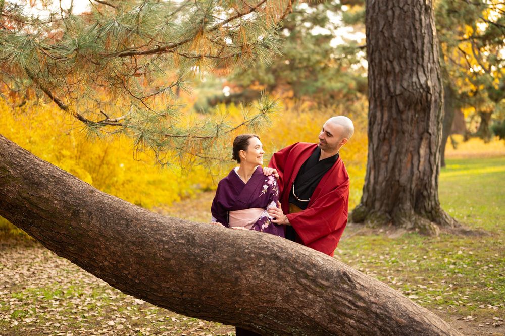 Couple in traditional Japanese kimono embracing beside a large leaning tree trunk in a park with golden autumn foliage.