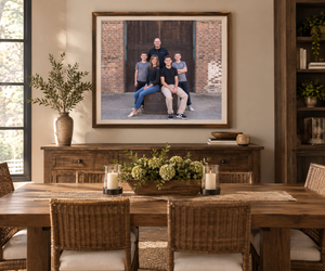 Rustic dining room with wooden table, wicker chairs, and a large framed family portrait above a sideboard with candles.
