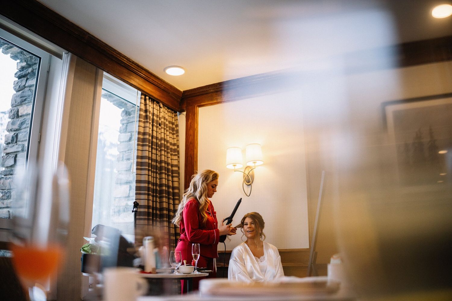 Bride in a white robe smiles as her hair is curled beside stone-framed windows before her Banff Christmas wedding