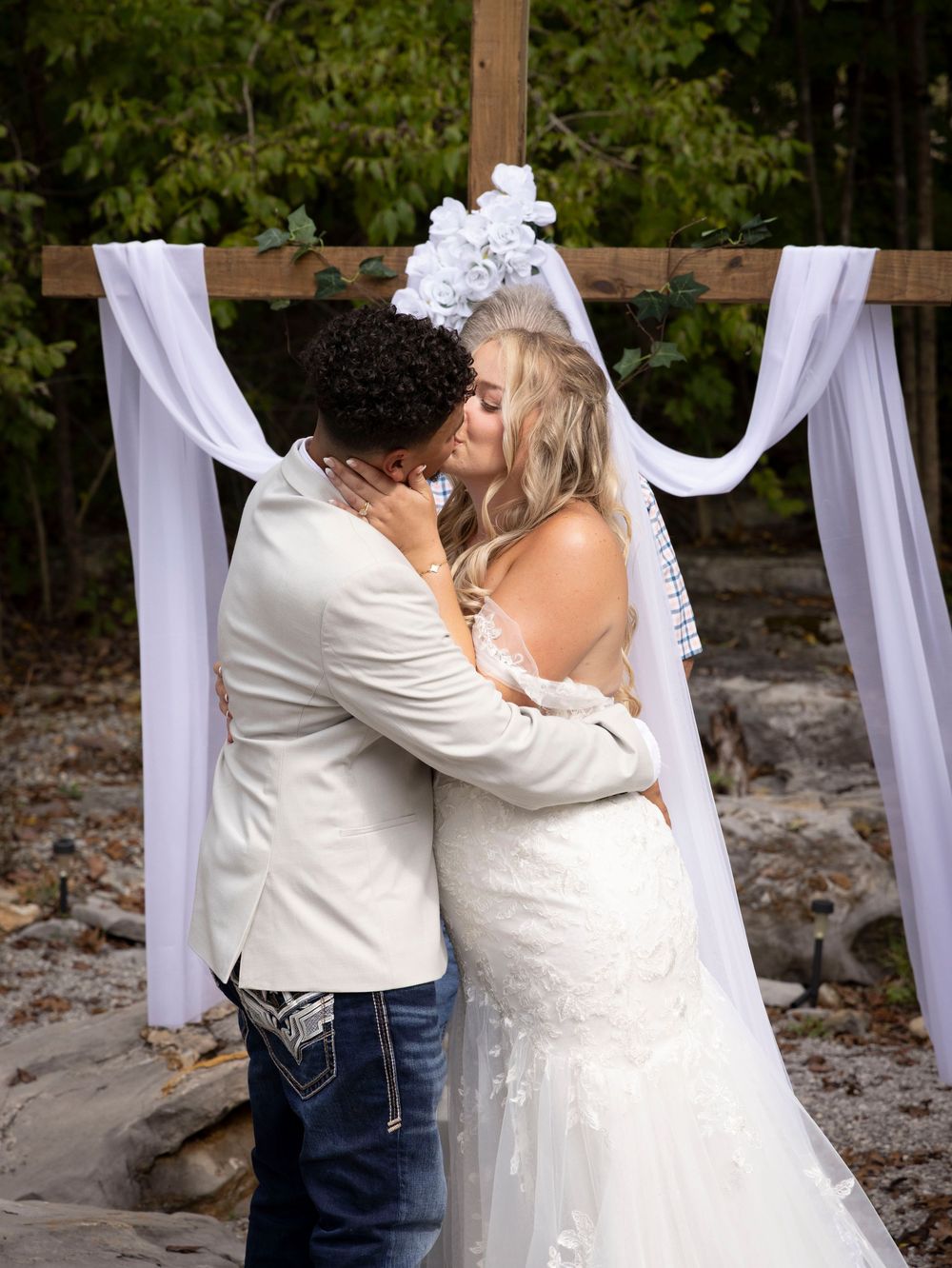 Couple sharing first kiss at outdoor wedding ceremony under white draped wooden arch surrounded by nature.