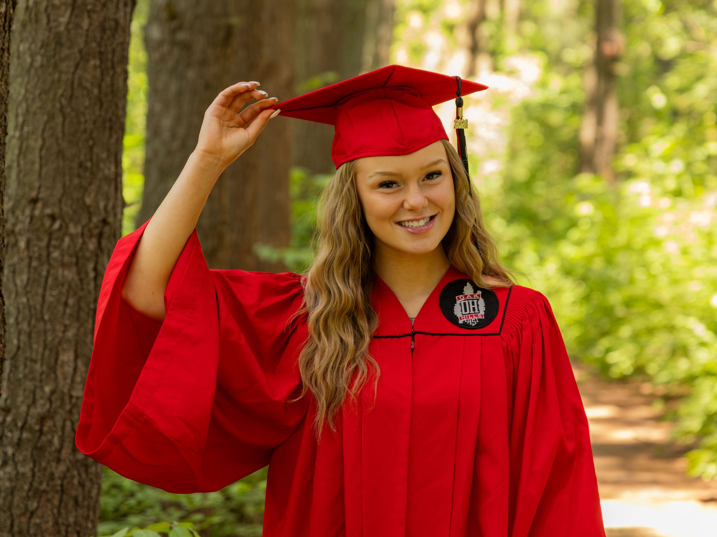 Young woman in red graduation cap and gown smiling outdoors among green trees, holding her cap.