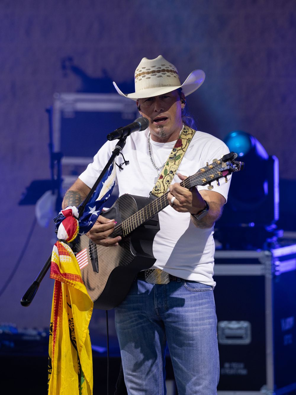 Country musician in white tee and cowboy hat plays acoustic guitar on stage near an American flag.