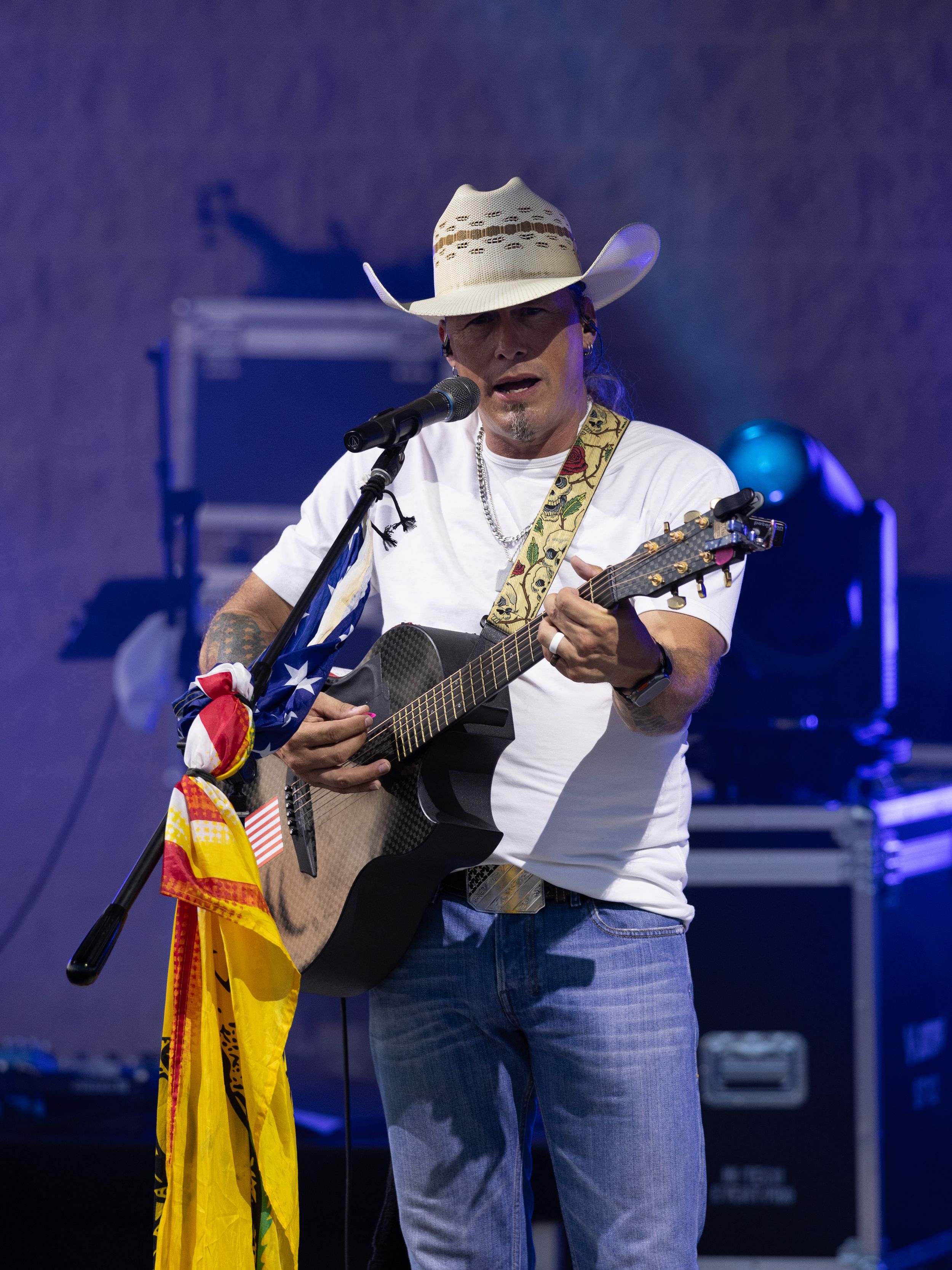 Country musician in white tee and cowboy hat plays acoustic guitar on stage near an American flag.