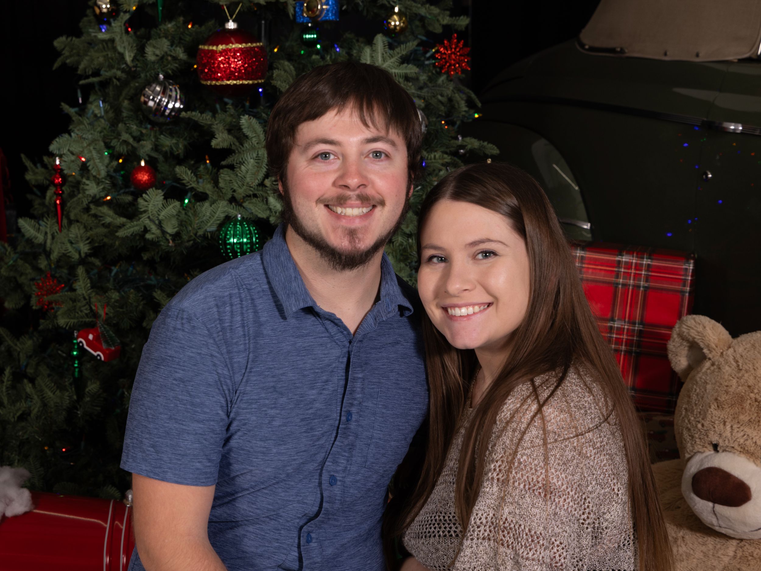 A smiling couple poses together in front of a decorated Christmas tree with gifts and a teddy bear nearby.