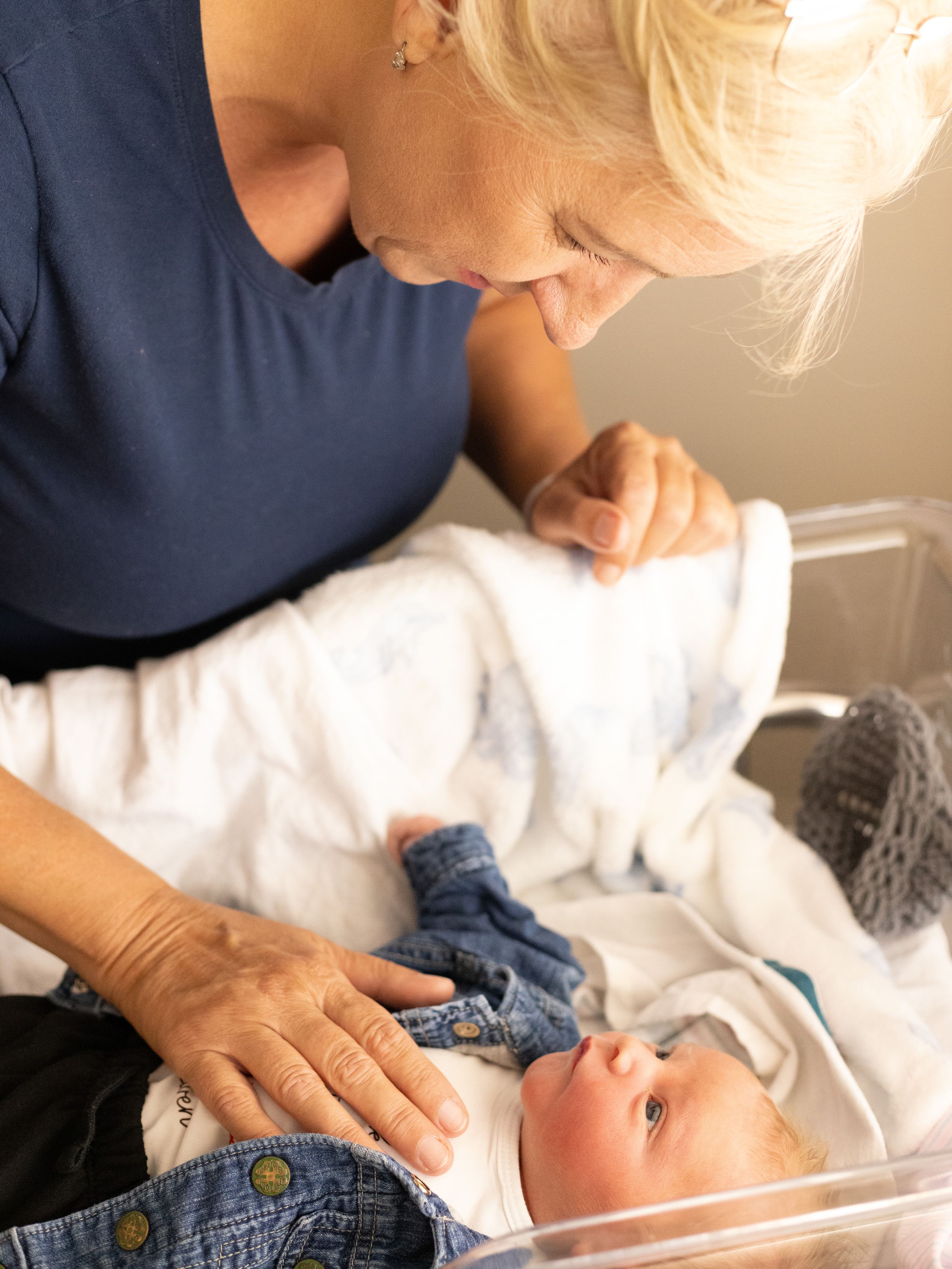Grandmother lovingly gazes at newborn baby lying in hospital bassinet, wearing denim outfit.