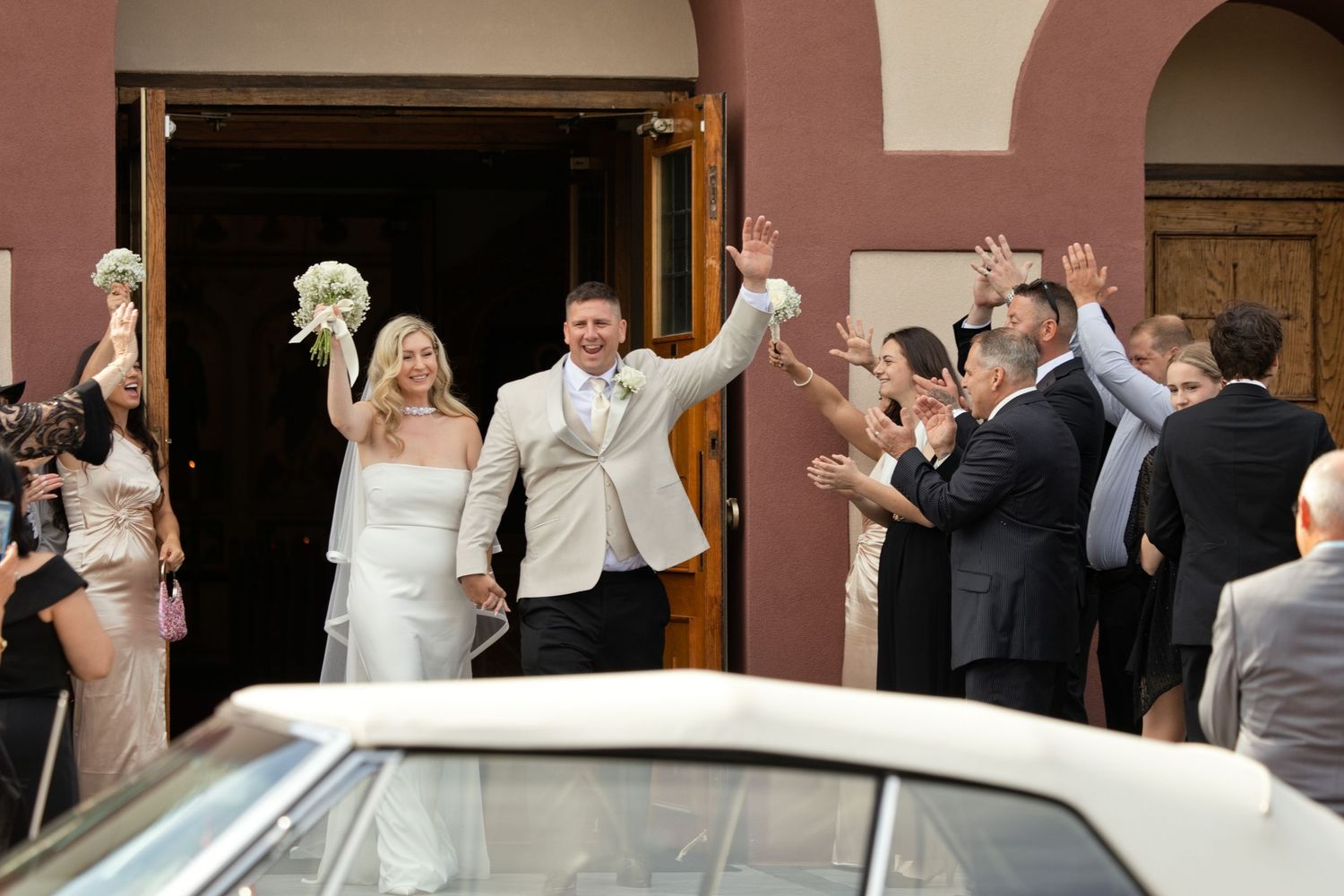 Newlyweds raise their arms in celebration outside a church as cheering wedding guests gather near a classic white car.