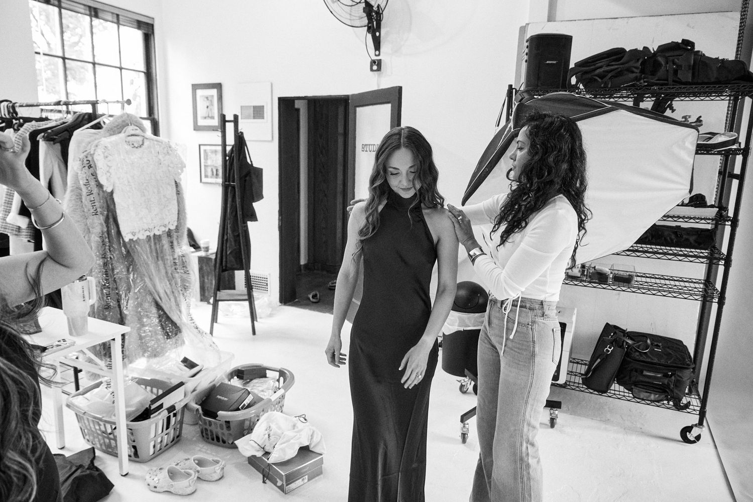 Smiling woman in wide-leg jeans and heels standing between hanging garments on a clothing rack in a bright studio.