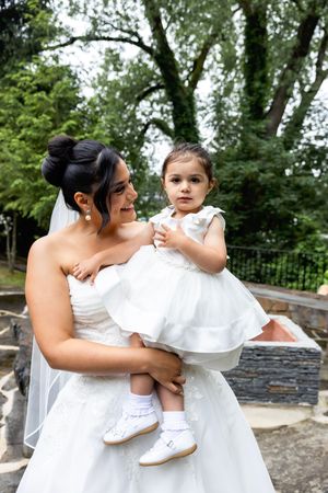 Bride in white gown and veil holds young flower girl in white dress at outdoor wedding venue.