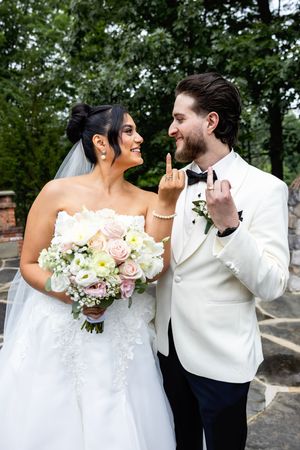 Smiling bride and groom in white tuxedo showing wedding rings outdoors, bride holding pink and white floral bouquet.