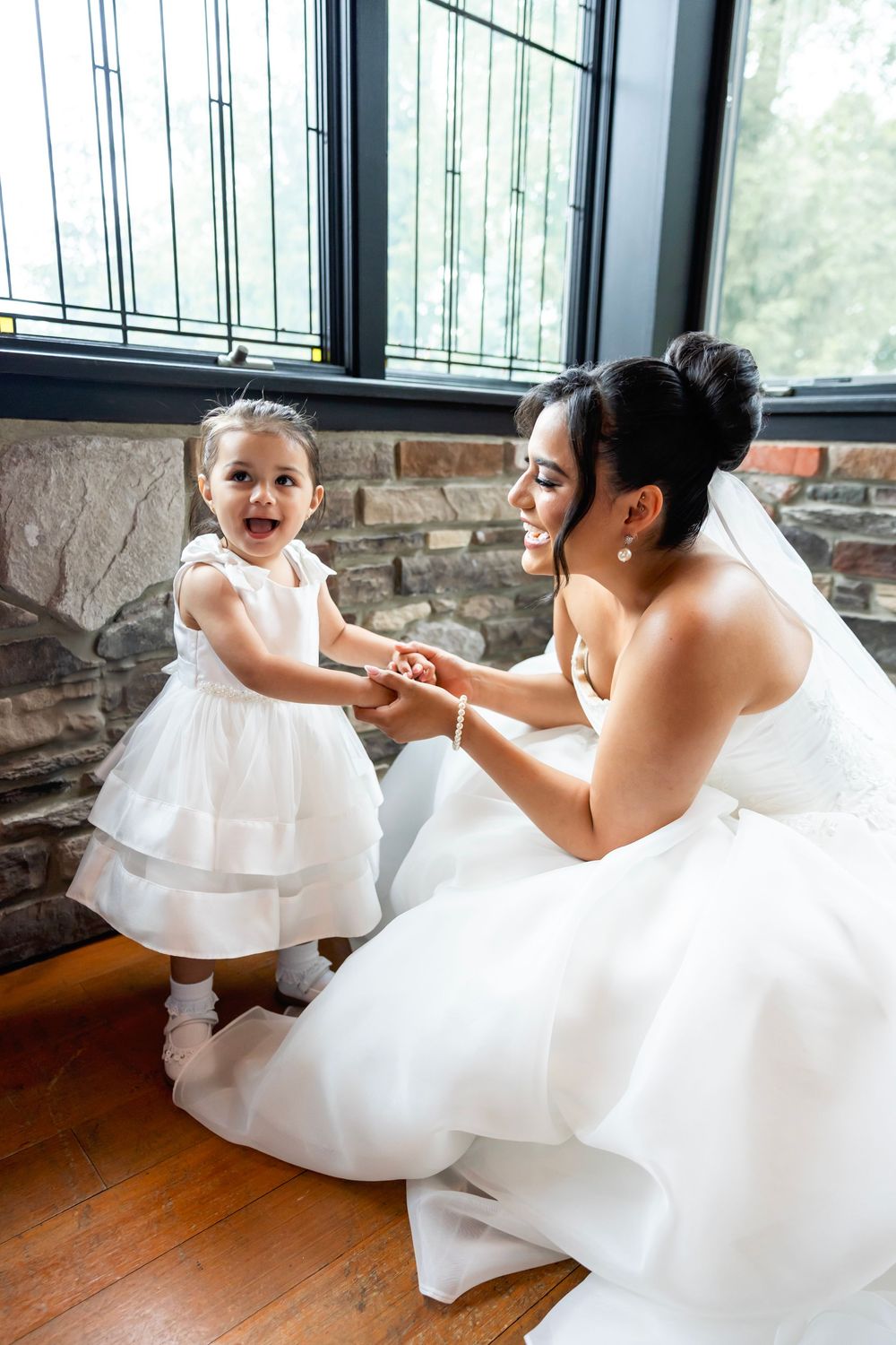 Bride in white gown kneels to hold hands with smiling flower girl in white dress near stone wall window.
