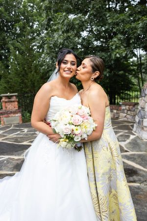 Bride in white strapless gown holding pink bouquet receives kiss on cheek from woman in yellow floral dress outdoors.