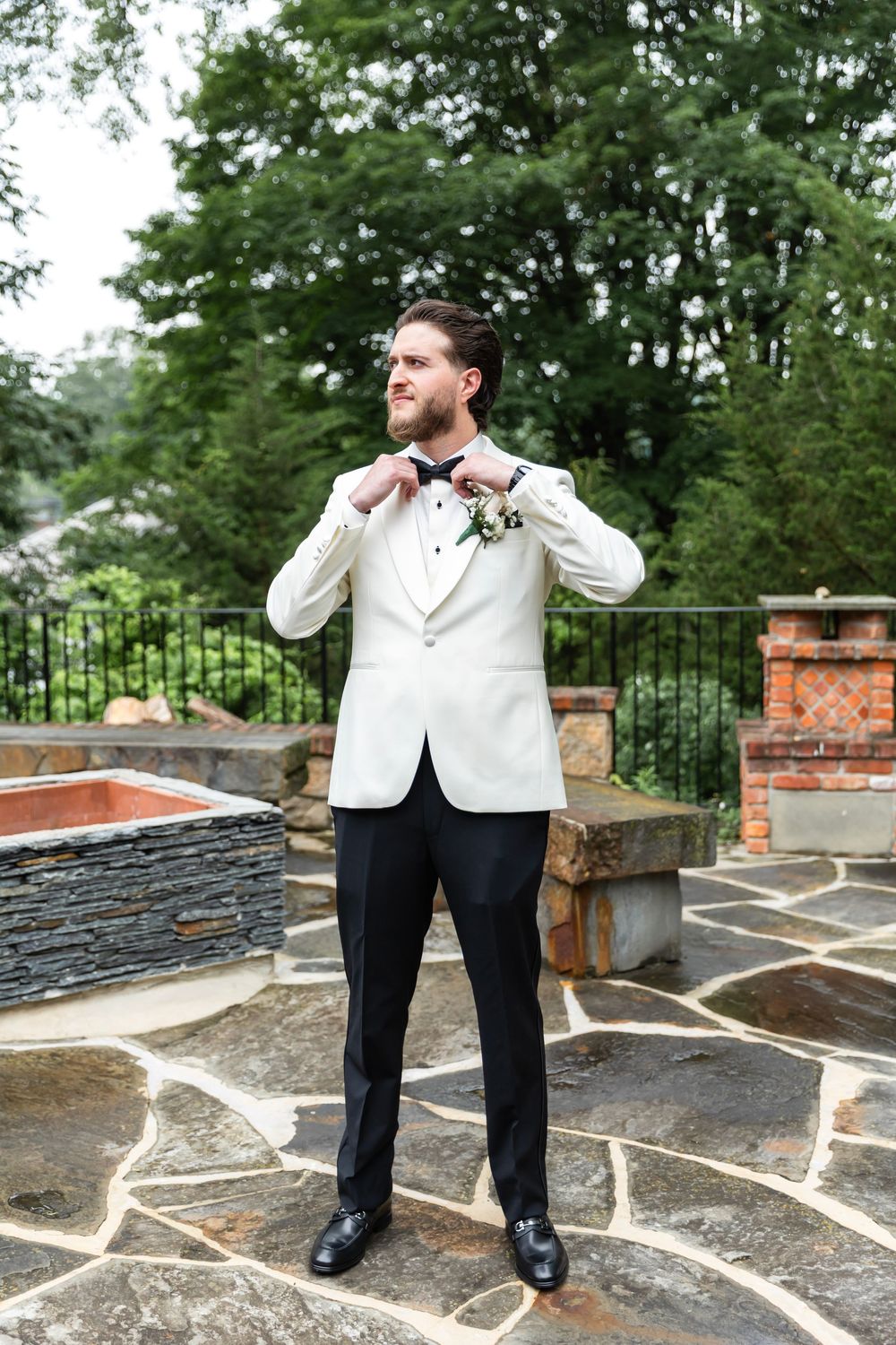 Groom in white tuxedo jacket adjusting bow tie on stone patio with lush green trees backdrop.