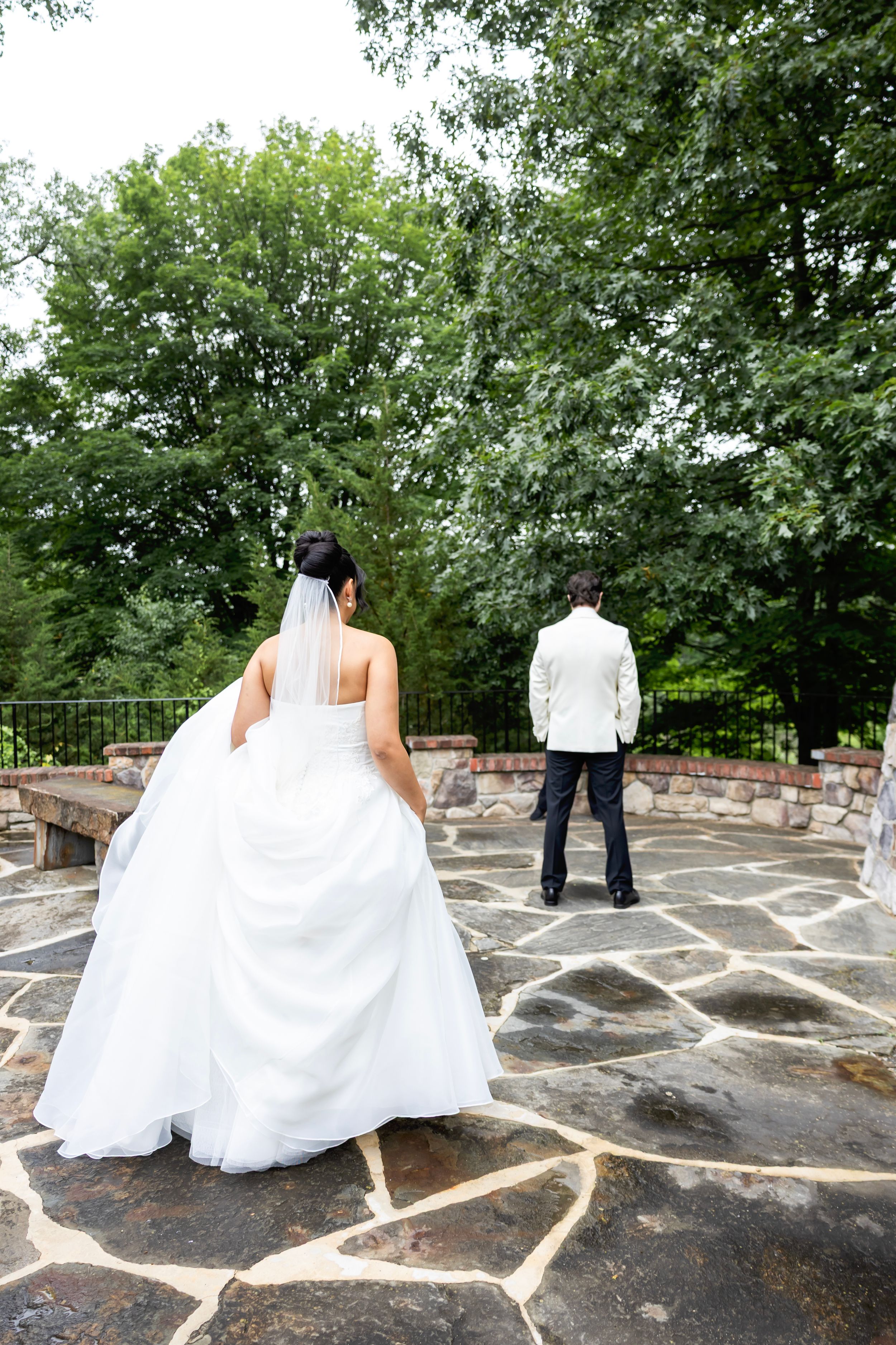 Bride in white gown and veil approaches groom in white suit on stone patio surrounded by lush green trees.