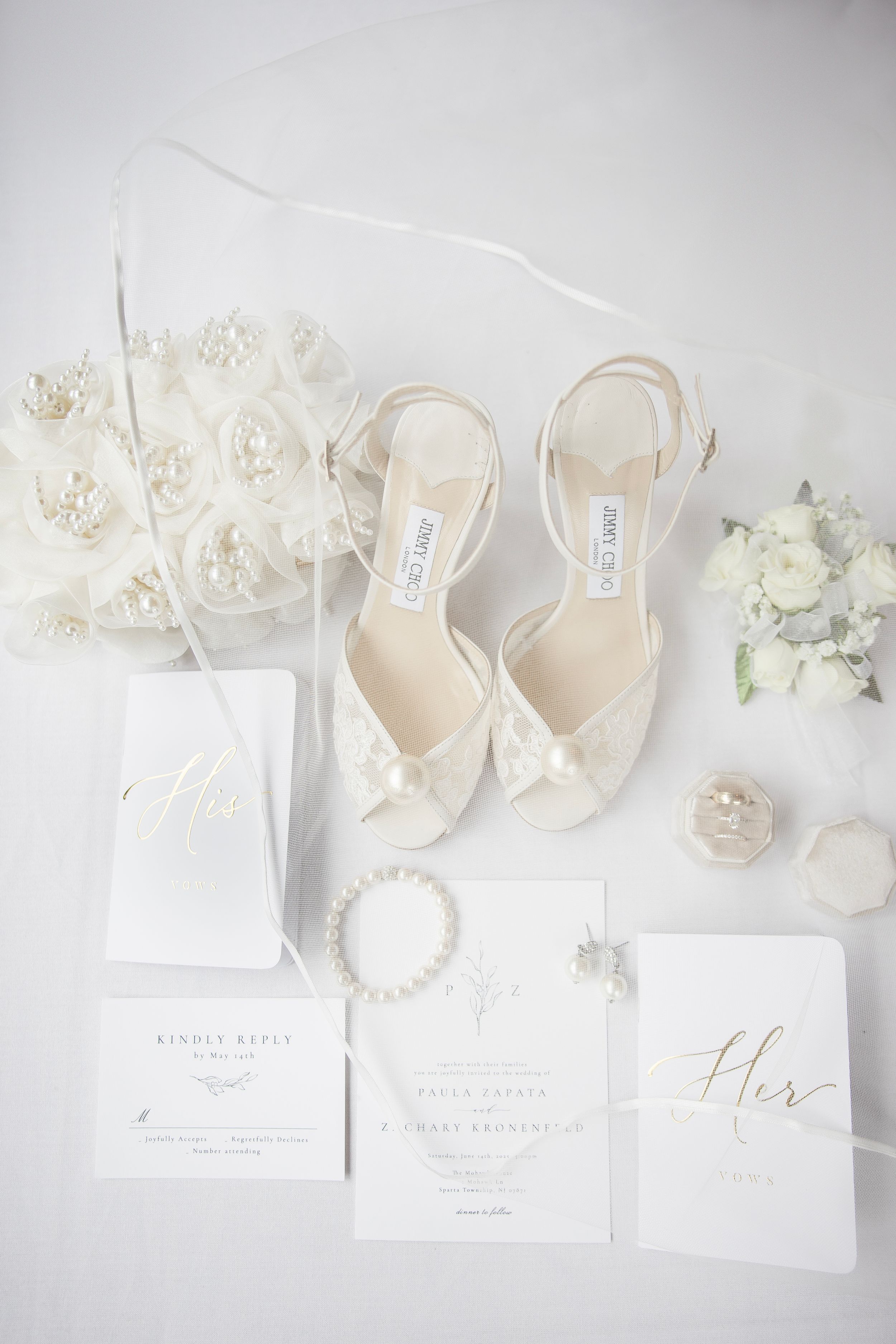 Elegant bridal flat lay featuring Jimmy Choo heels, pearl bracelet, white floral bouquet, and wedding stationery on white background.