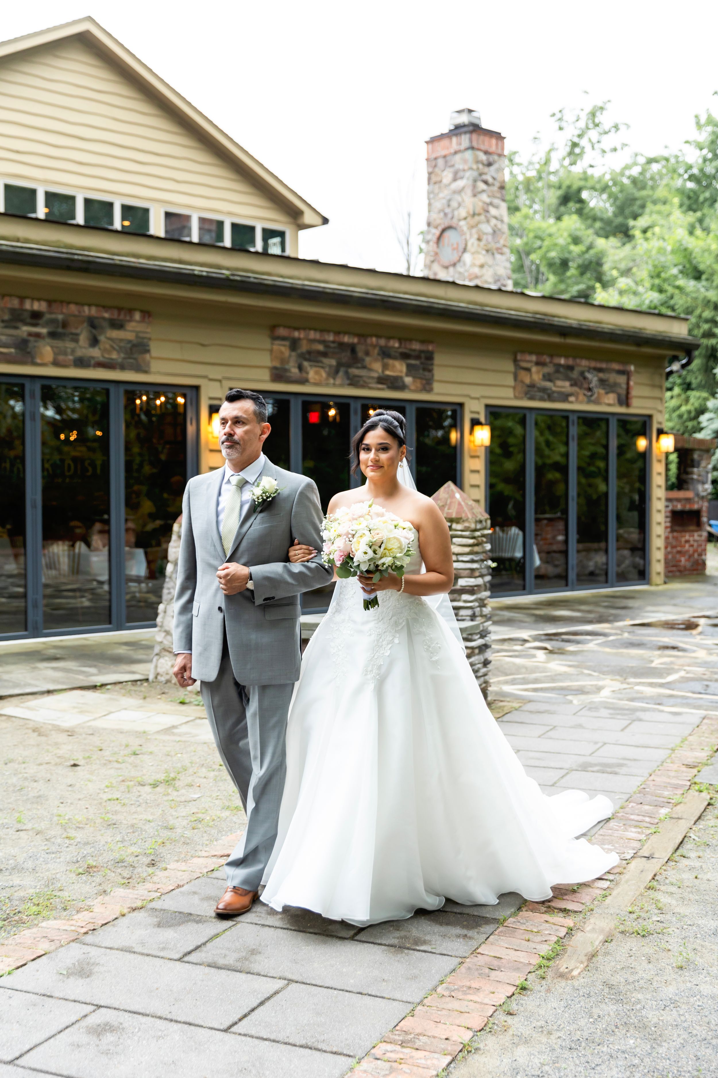 Bride in white gown and groom in gray suit walk together on stone path outside rustic venue on wedding day.