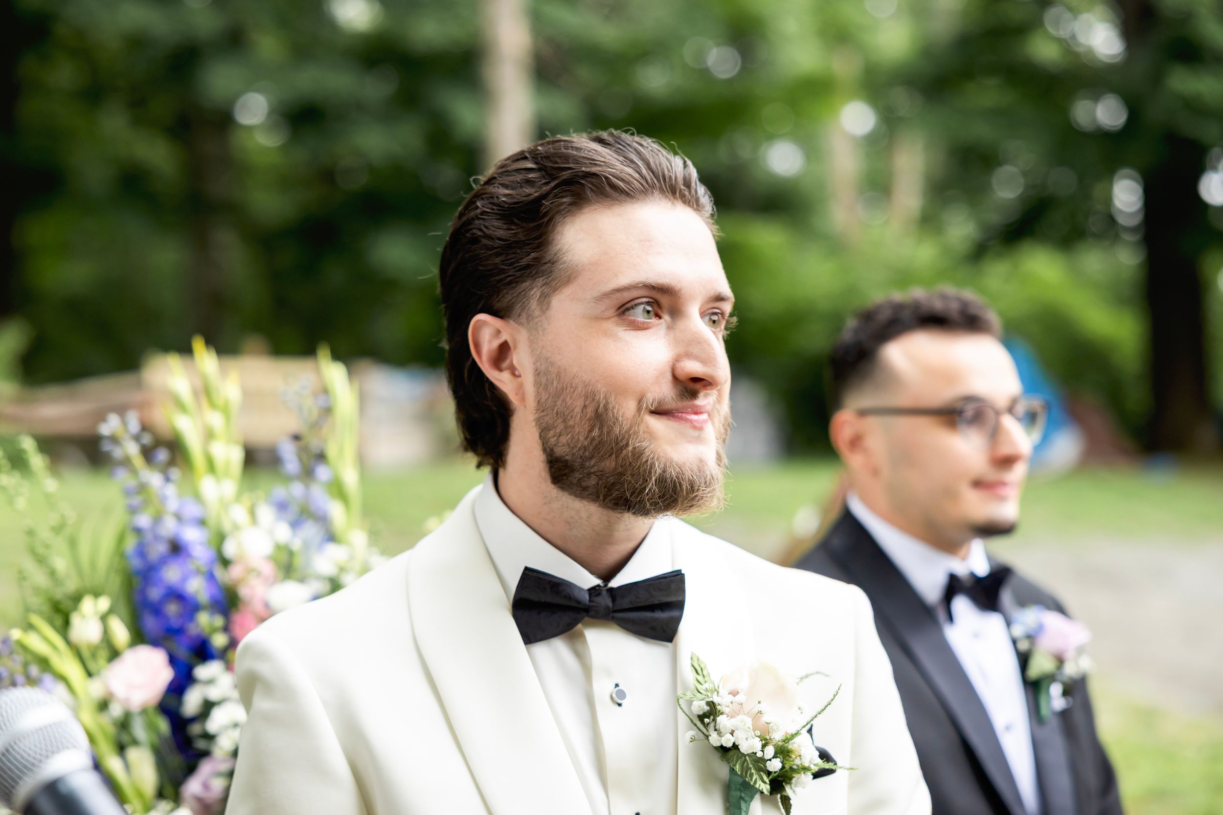 Groom in white tuxedo with bow tie and boutonniere smiling at wedding ceremony outdoors.