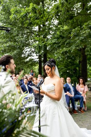 Bride reads wedding vows at outdoor ceremony surrounded by lush green trees and seated guests.