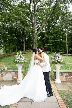 Bride and groom share a kiss at outdoor wedding ceremony with floral arrangements on white columns and lush green trees.
