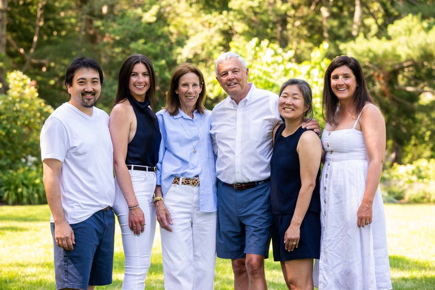 Six smiling family members pose together outdoors on a sunny day in a lush green garden setting.