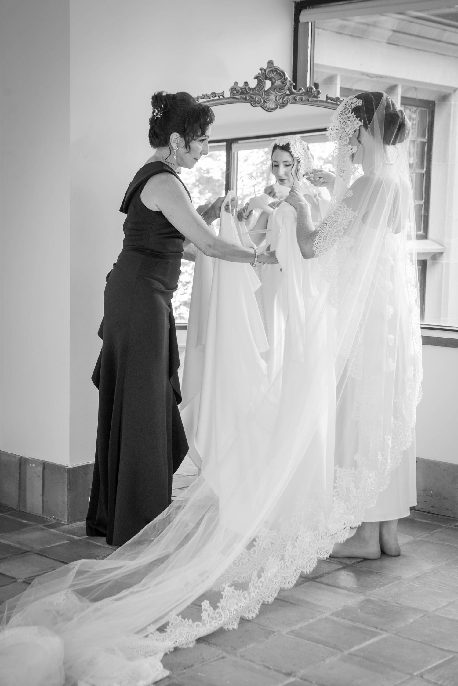 Black and white photo of a bride in a lace gown and veil being helped by a woman in a black dress before her wedding.