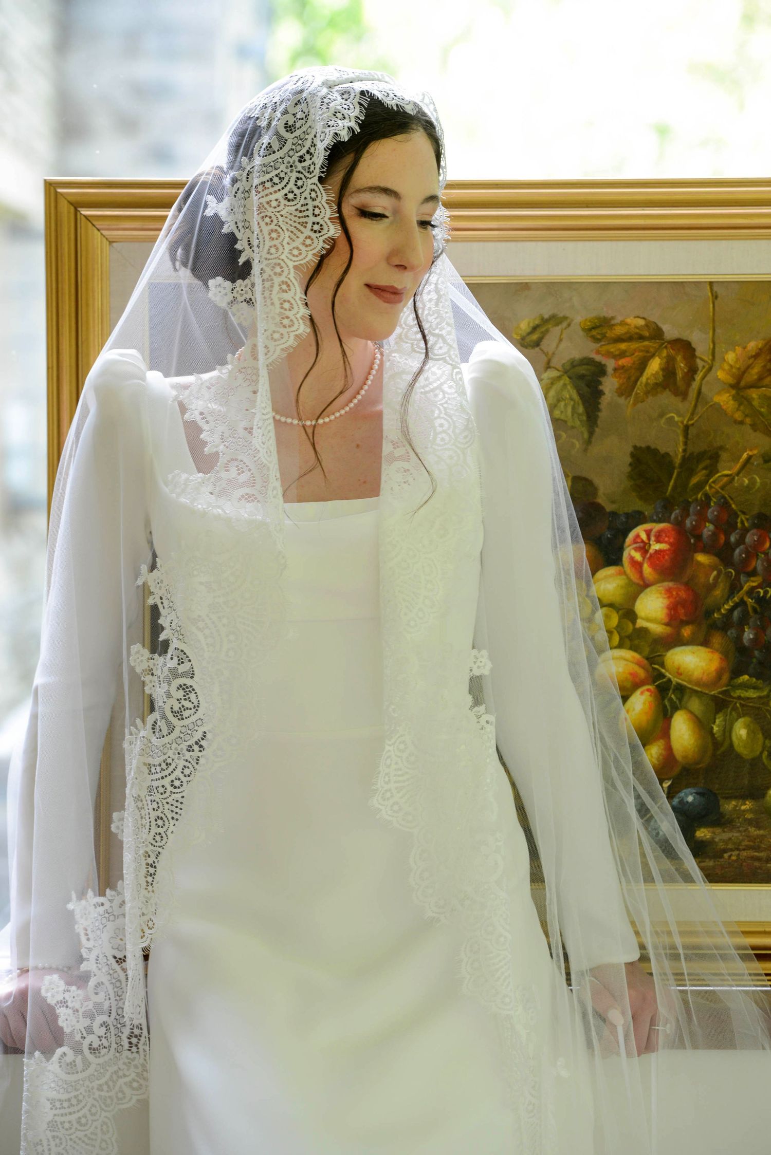 Bride in white lace wedding dress and veil poses near a fruit painting in natural light.