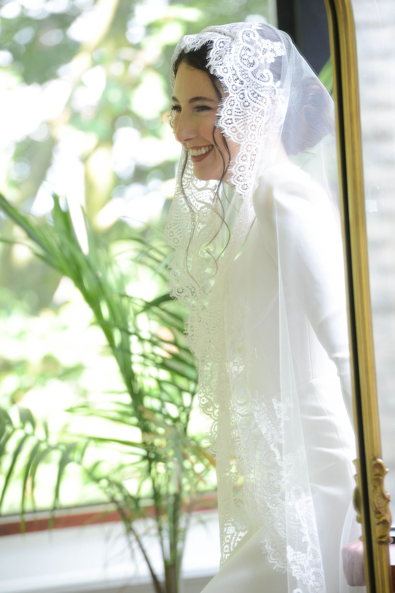Smiling bride in white lace veil and gown standing by a sunlit window with tropical plants in background.