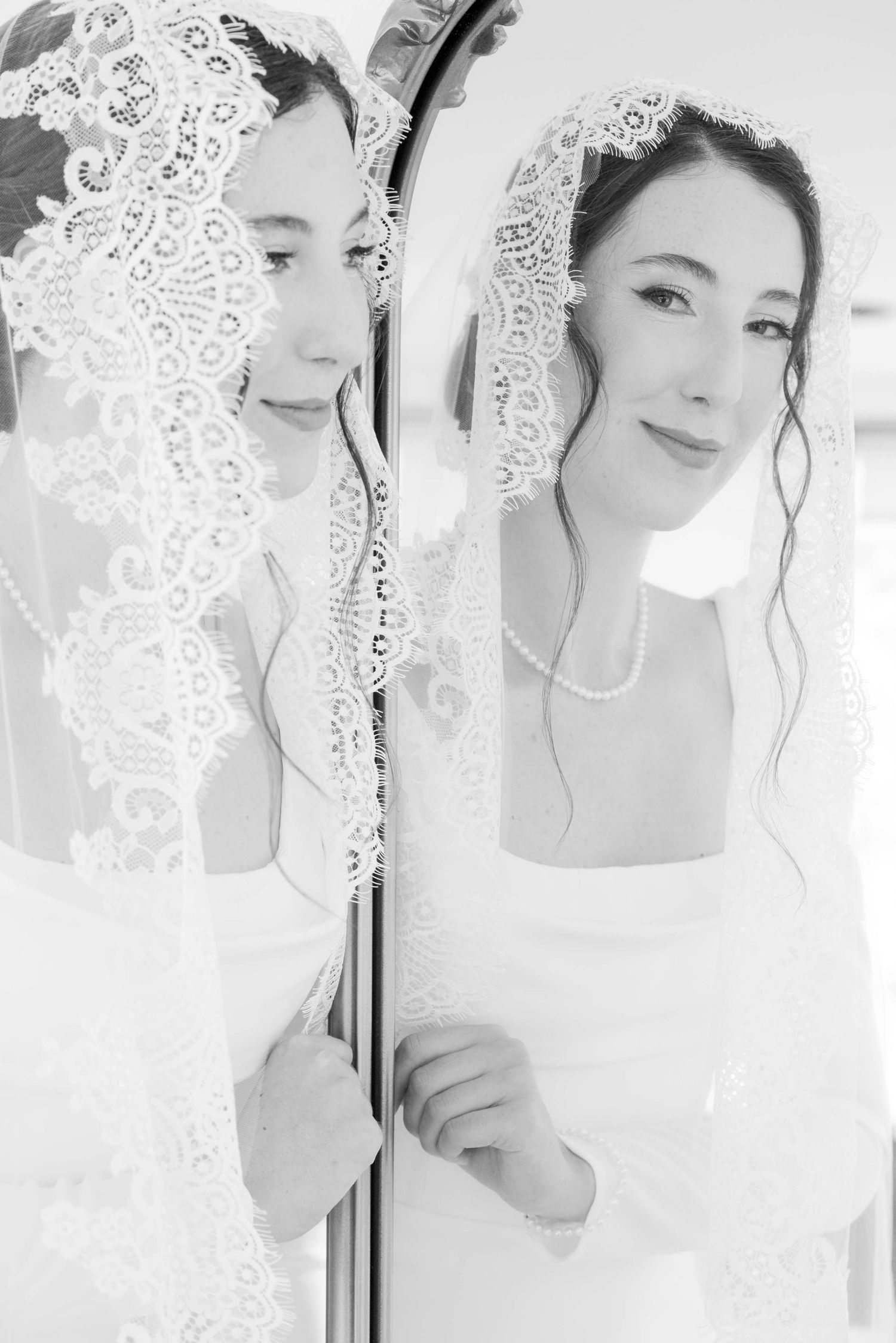Black and white photo of a bride in lace veil and pearl necklace gazing at her reflection in a mirror.