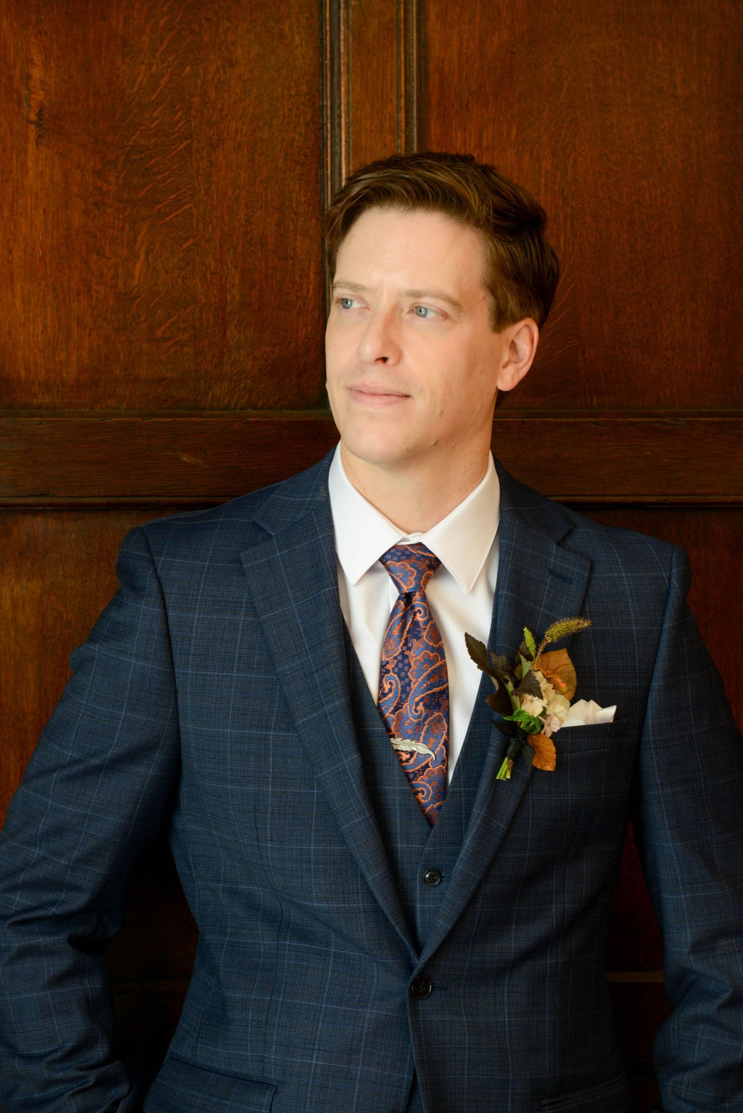 Groom in navy plaid suit with floral boutonniere and paisley tie posing against wood paneling.