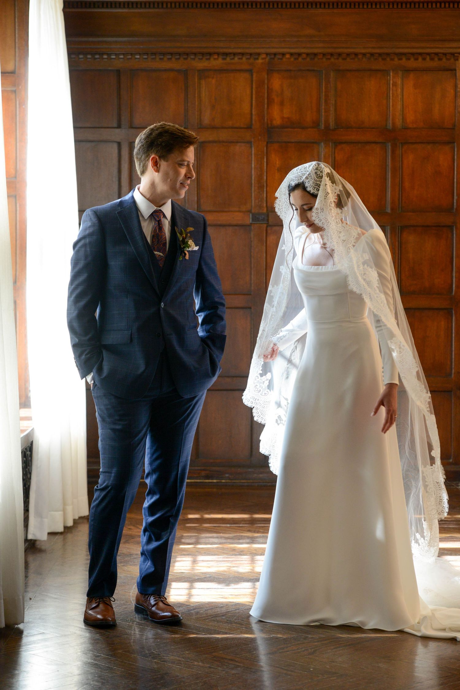 Bride in white gown with lace veil and groom in navy suit share first look moment indoors.
