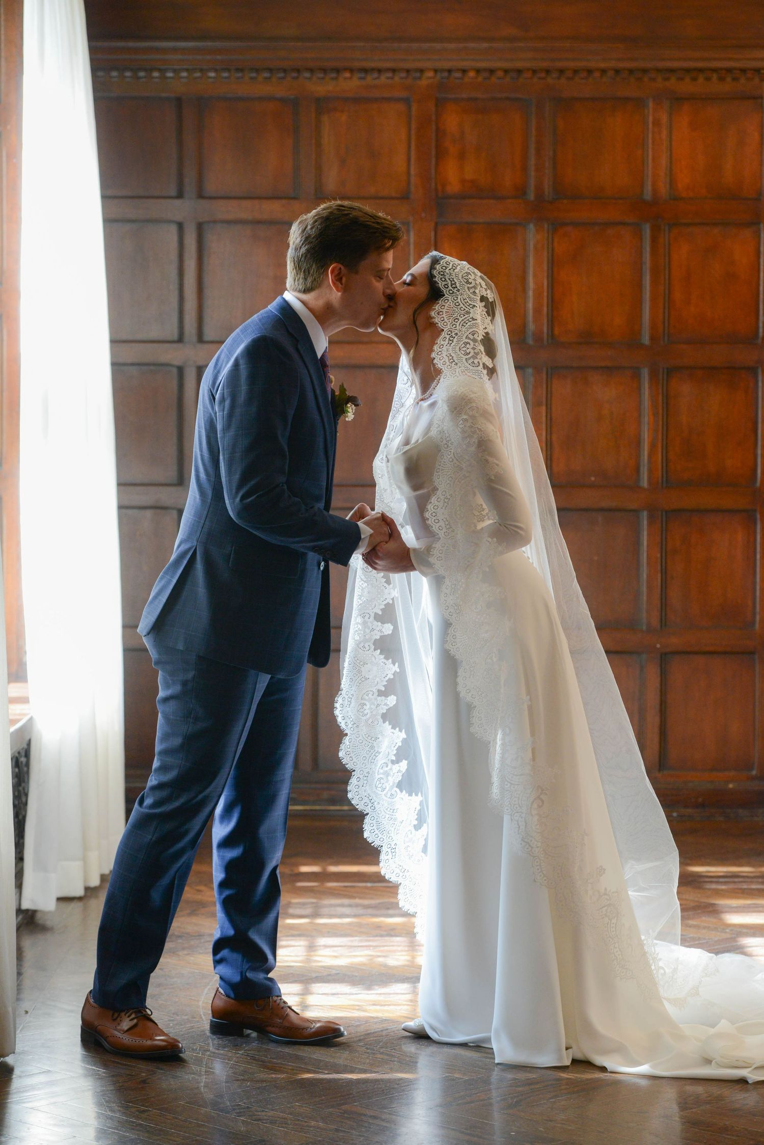 Bride and groom share a first kiss in elegant wood-paneled room, bride in lace veil and gown, groom in blue suit.