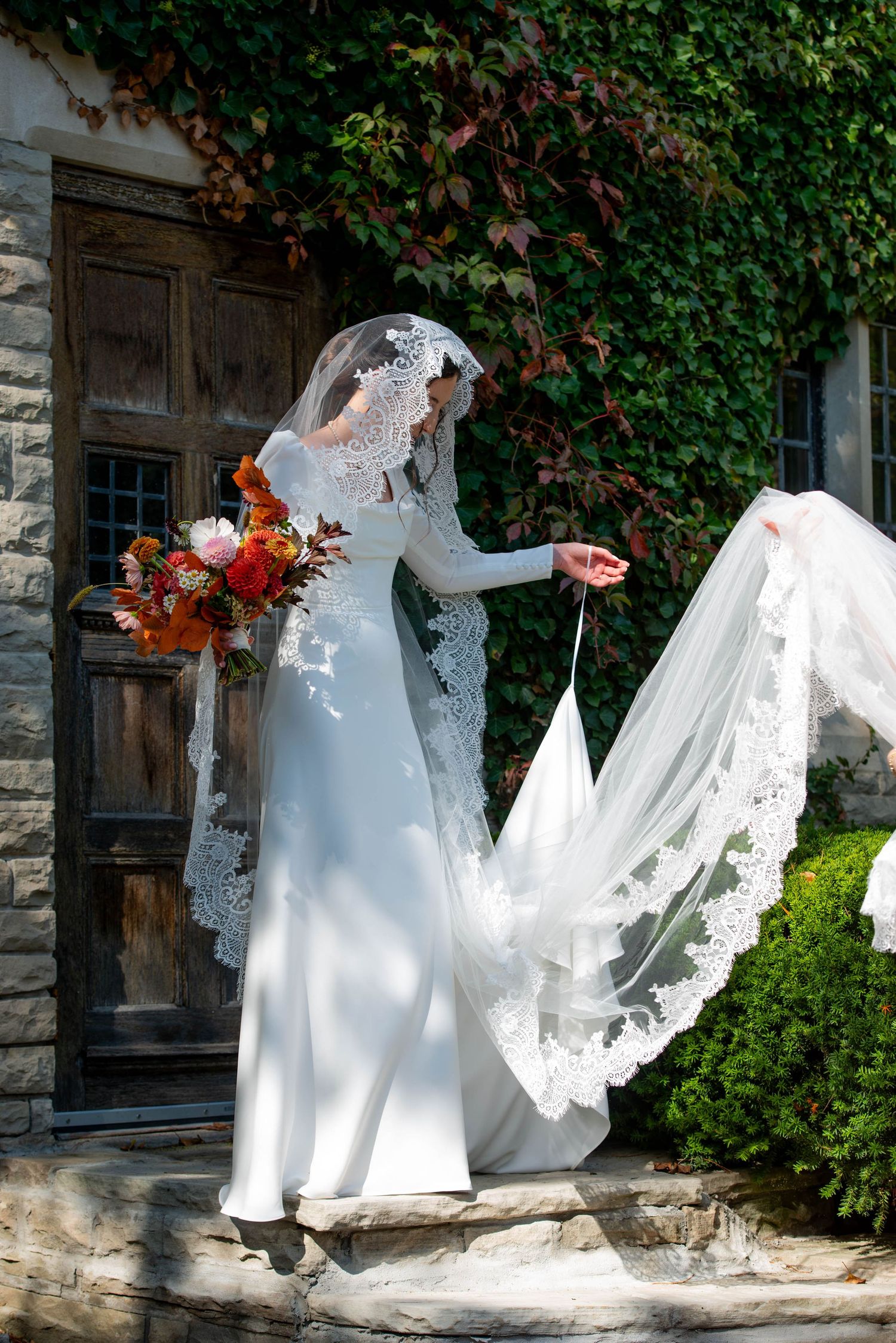 Bride in white lace wedding gown with flowing veil and autumn bouquet stands on stone steps by ivy-covered wall.