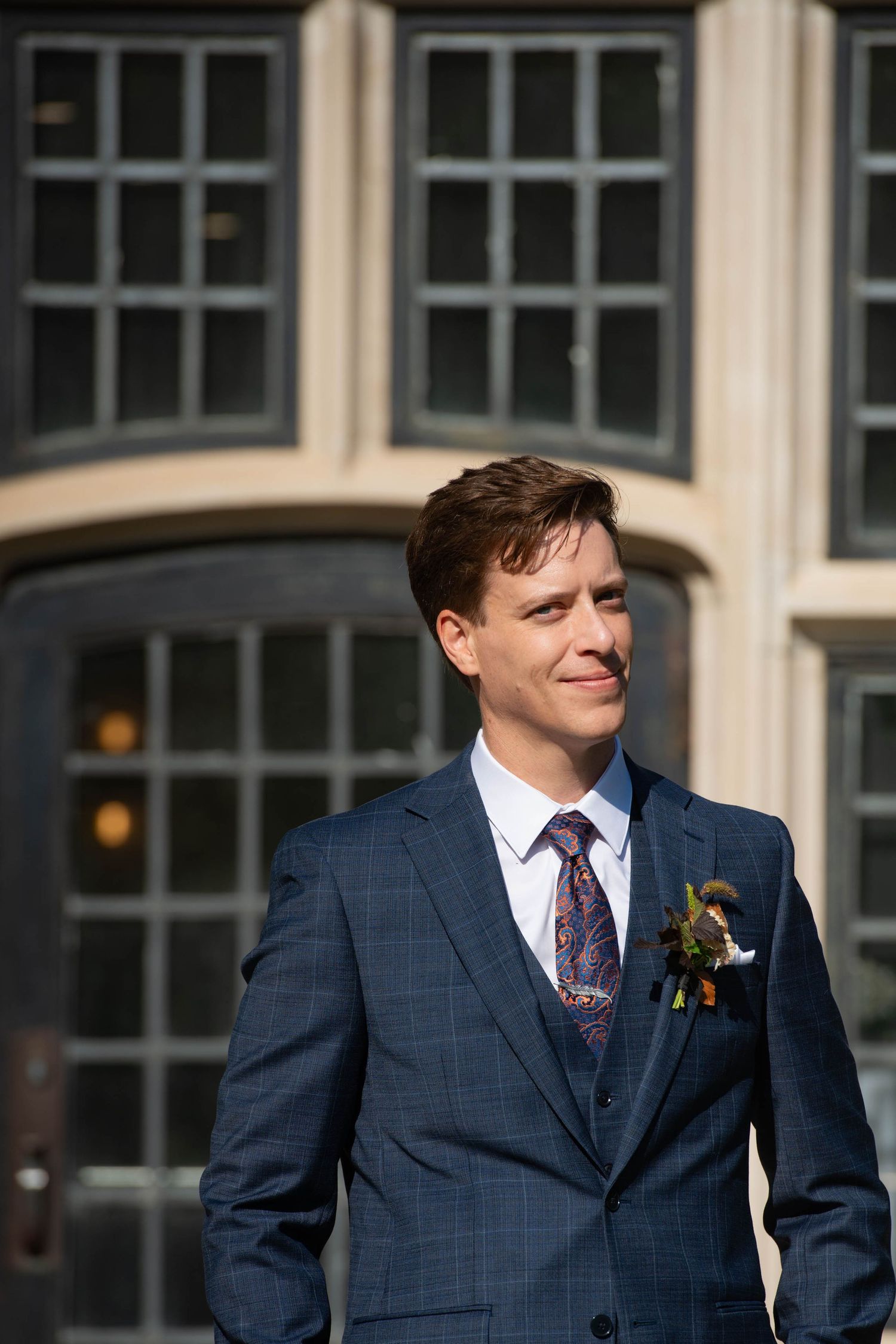 Groom in navy blue suit with floral boutonniere smiling outdoors before elegant building backdrop.