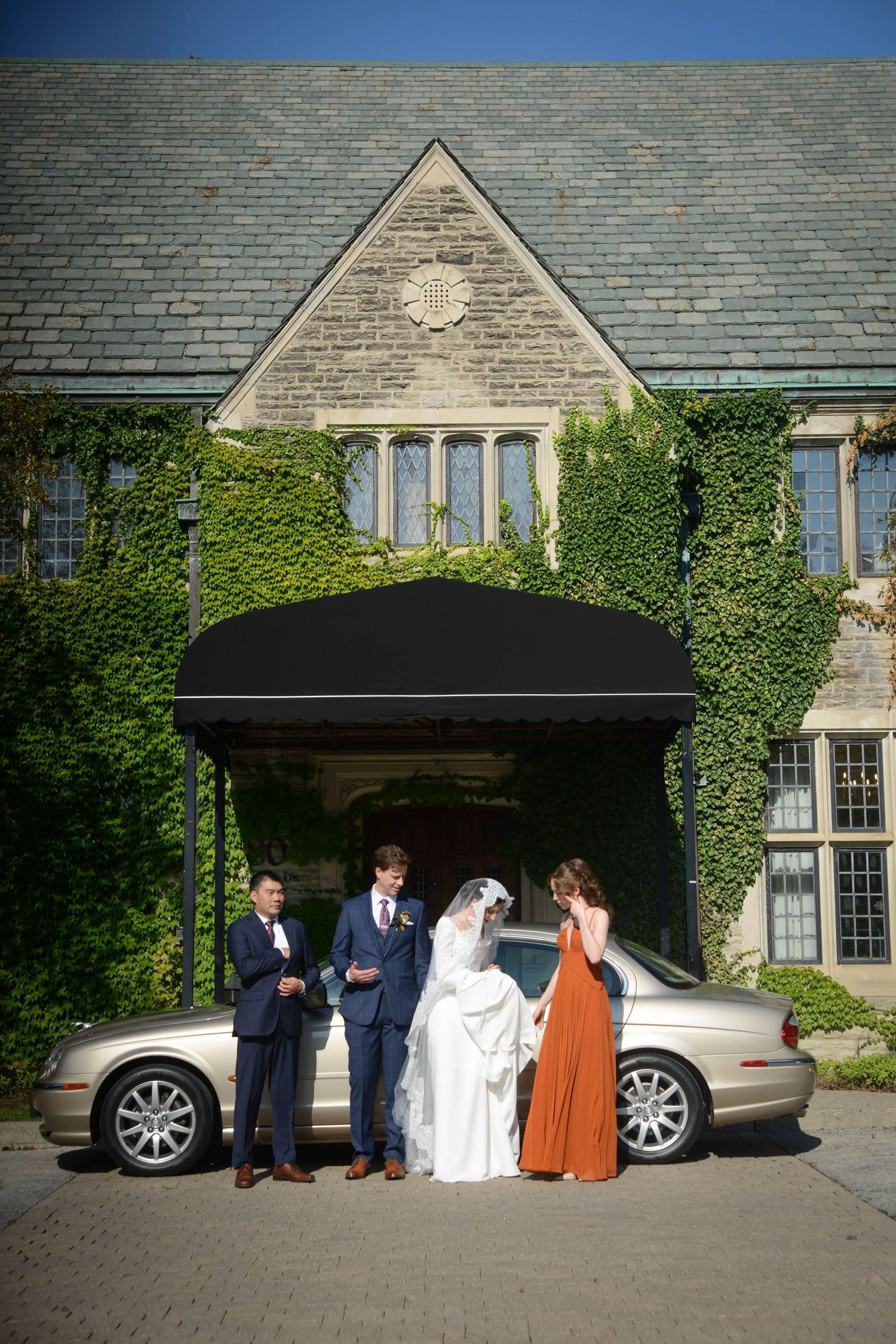 Wedding party poses by a vintage car outside an ivy-covered stone mansion on a sunny day.