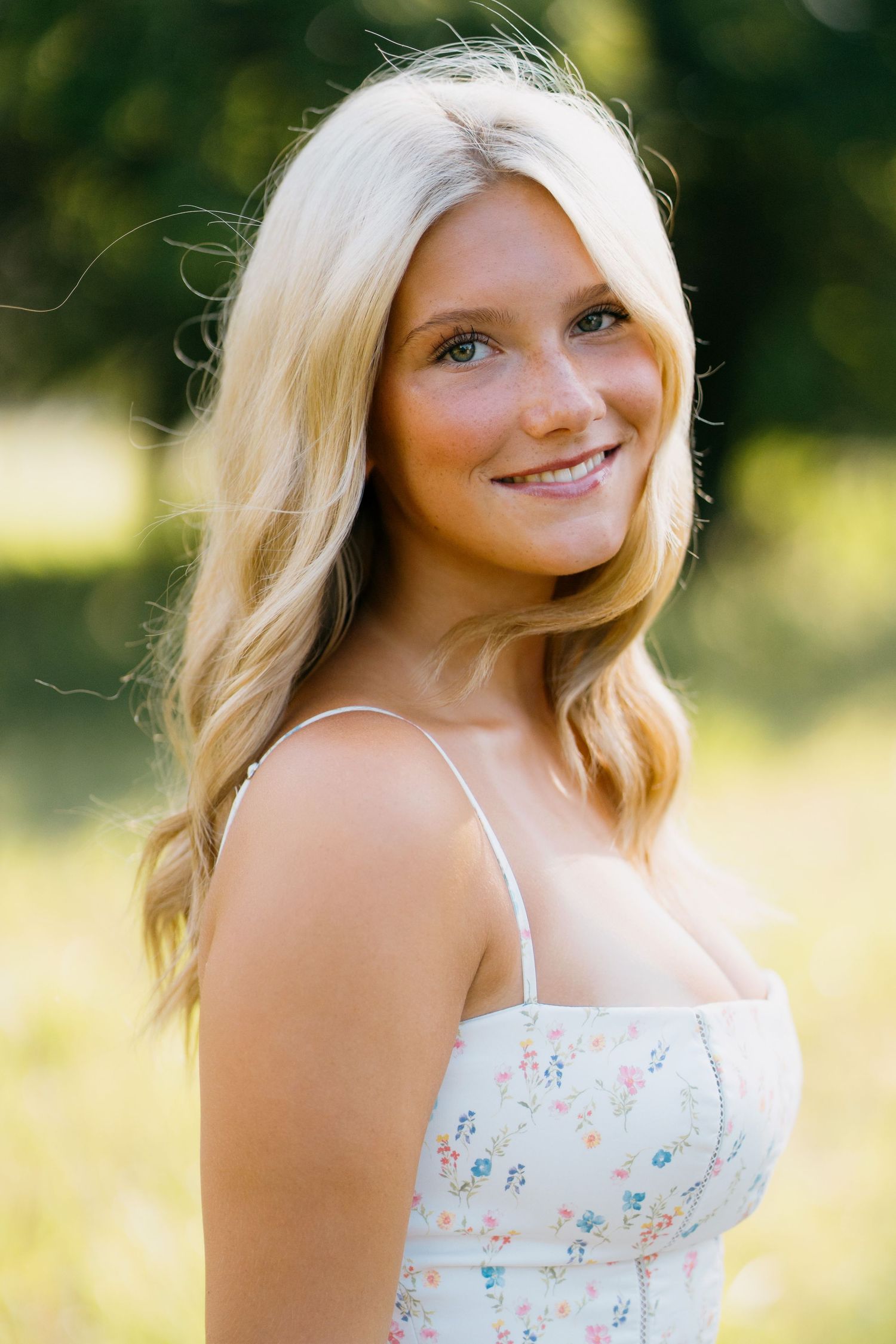 Smiling young blonde woman in floral sundress posing outdoors in a sunlit green park setting.