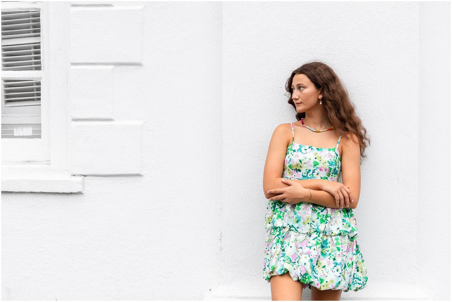 Young woman in floral sundress leaning against white wall outdoors.