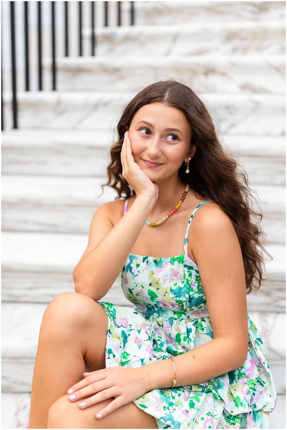 Young woman in floral dress sitting on marble steps, smiling dreamily.