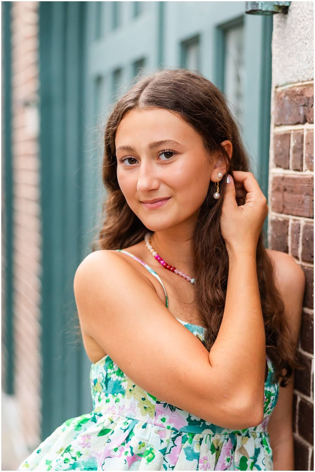Young woman in floral dress leaning against brick wall, touching her pearl earring and smiling softly.