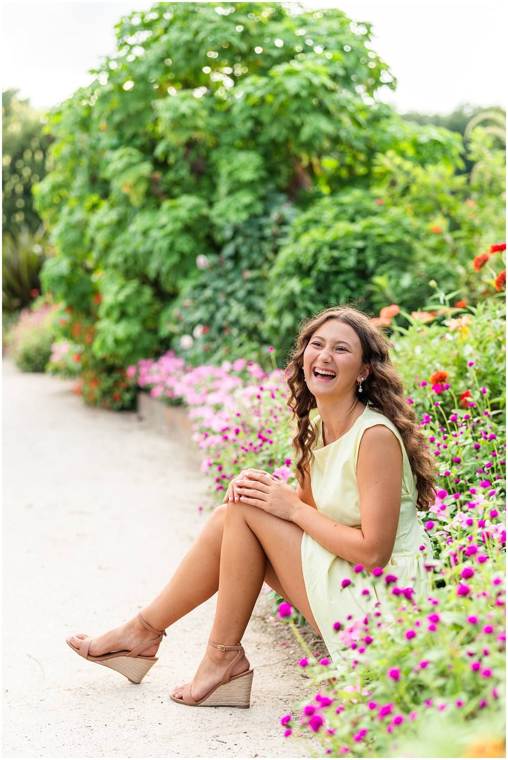 Laughing young woman in mint dress sitting among colorful garden flowers for senior portrait session.