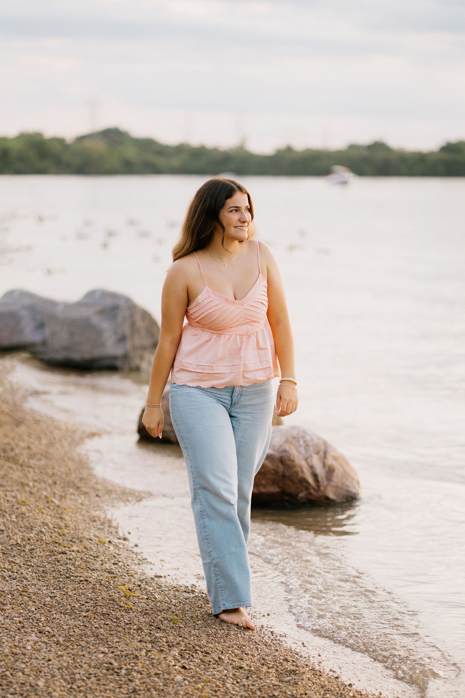 Young woman in pink tank top and jeans walking barefoot along a rocky shoreline with calm water in the background.