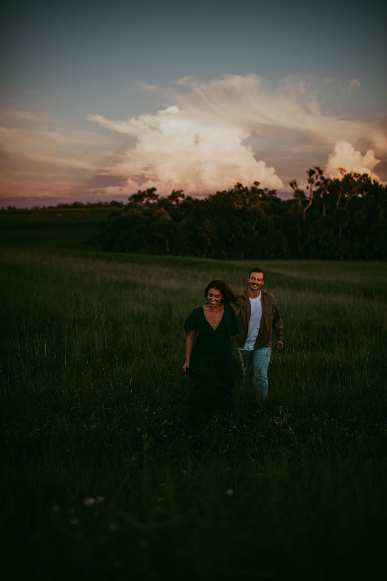 Couple walking hand in hand through lush green field at sunset with dramatic pink clouds in background.