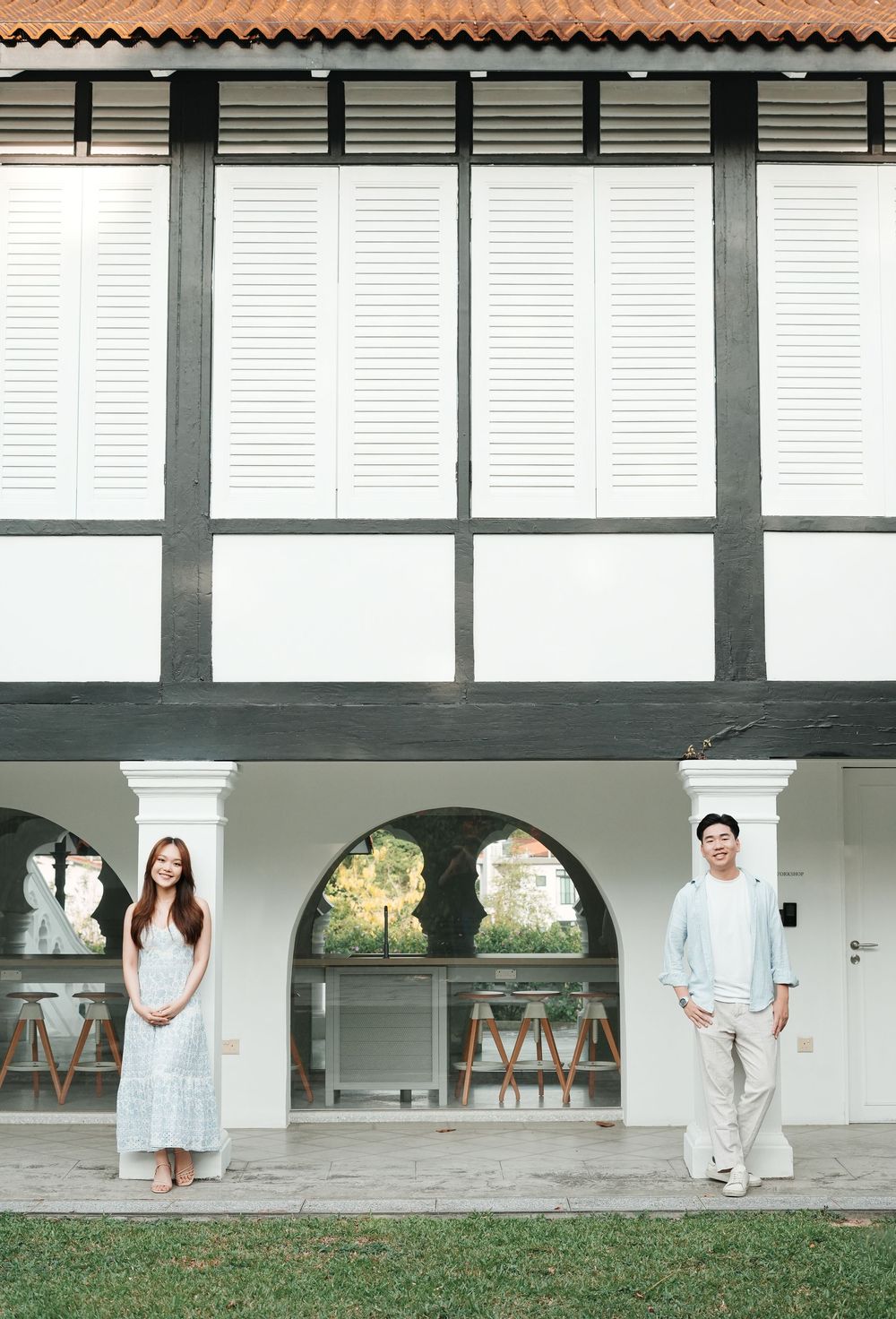Couple in light outfits standing apart under white arches of a colonial building exterior.