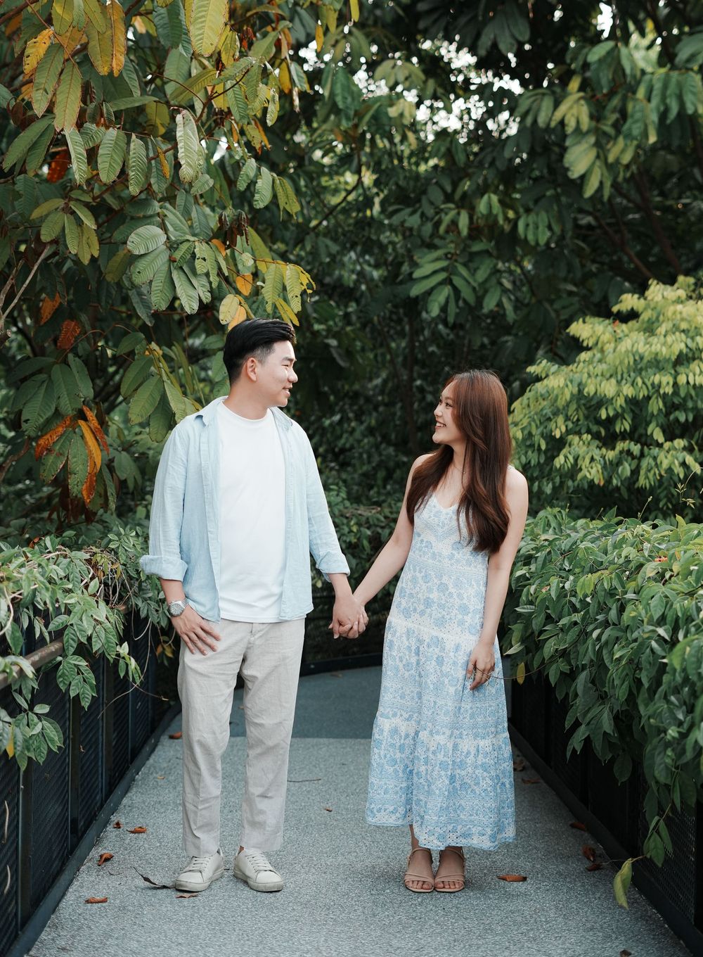 A couple holds hands and smiles at each other on a garden path surrounded by lush green trees.