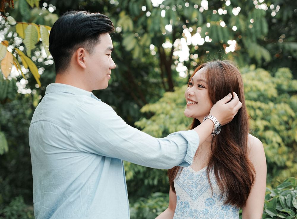 A young Asian couple shares a tender moment outdoors, as the man gently fixes the woman's hair while she smiles.