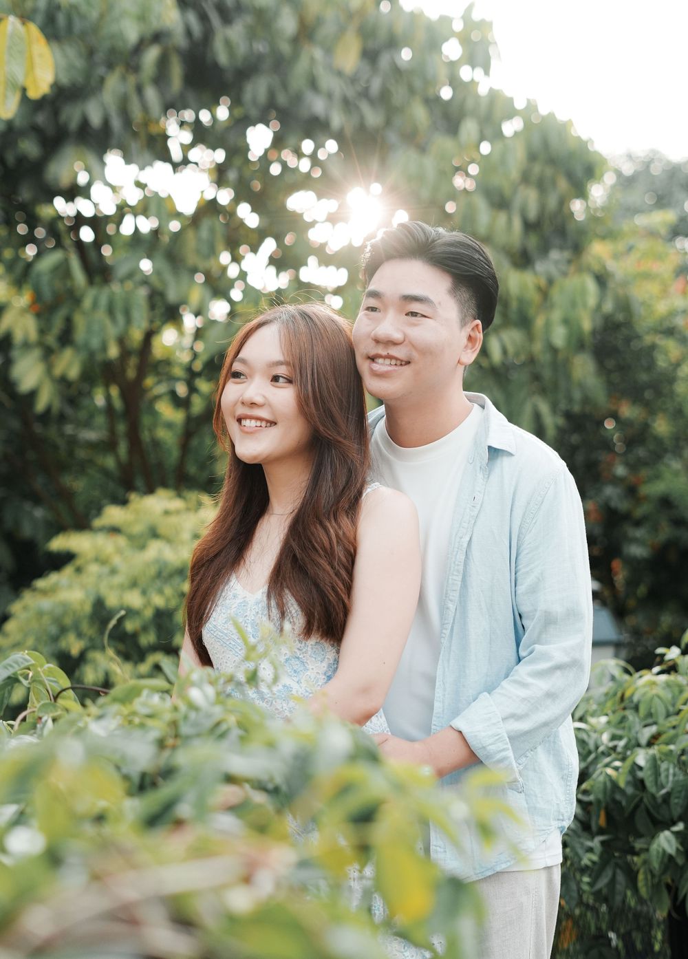 A smiling Asian couple poses together in a lush green garden with sunlight filtering through the trees.