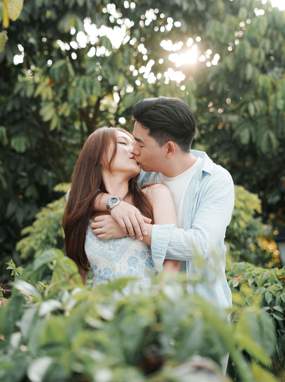 A couple shares a romantic kiss surrounded by lush green foliage with golden sunlight filtering through trees.
